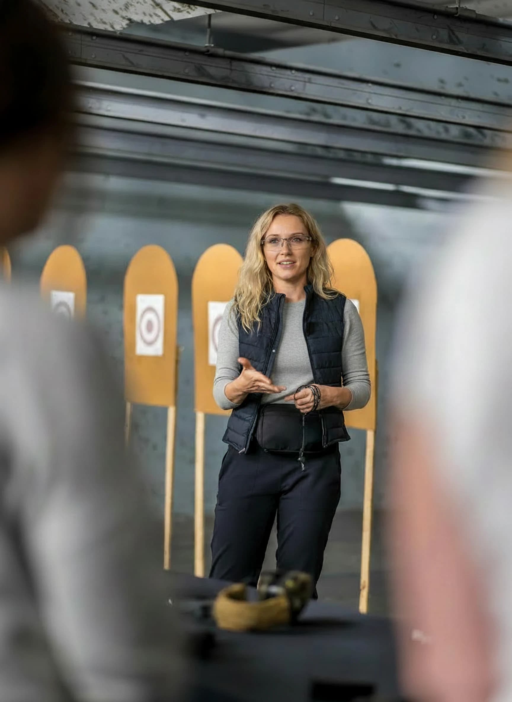 A GFT instructor in a branded vest explaining a point to learners on an indoor shooting range